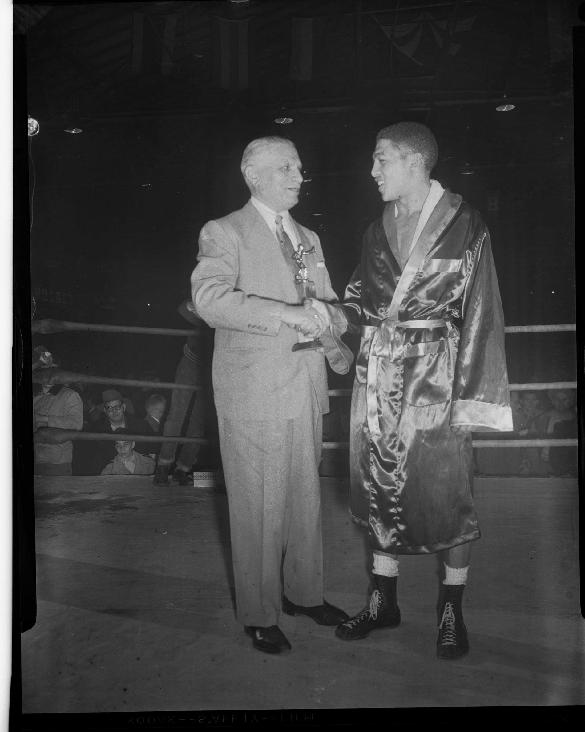 Boxer, possibly Bob Baker, in robe receiving trophy from man in ring in
