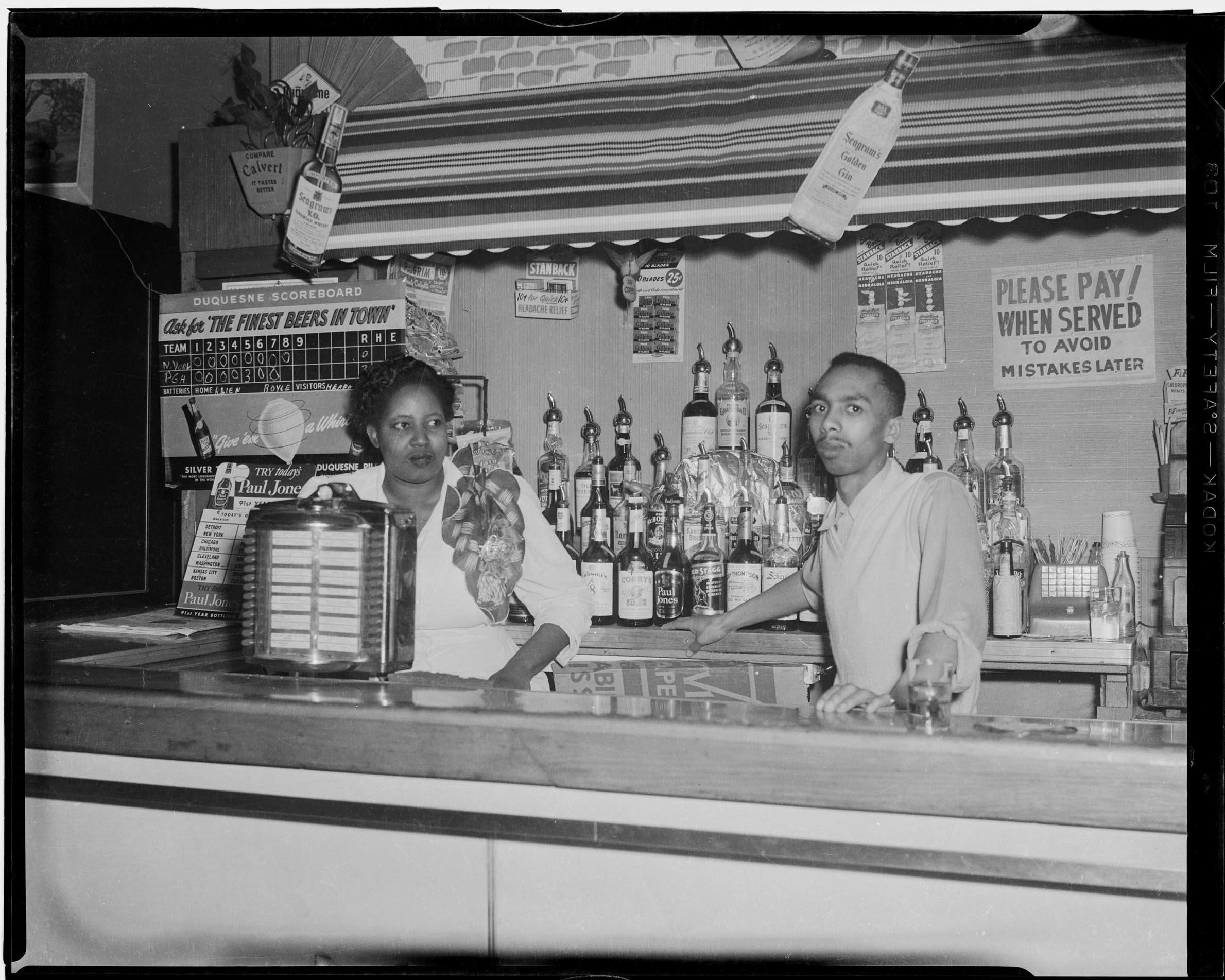 Woman and man behind bar, with Duquesne Beer scoreboard in background