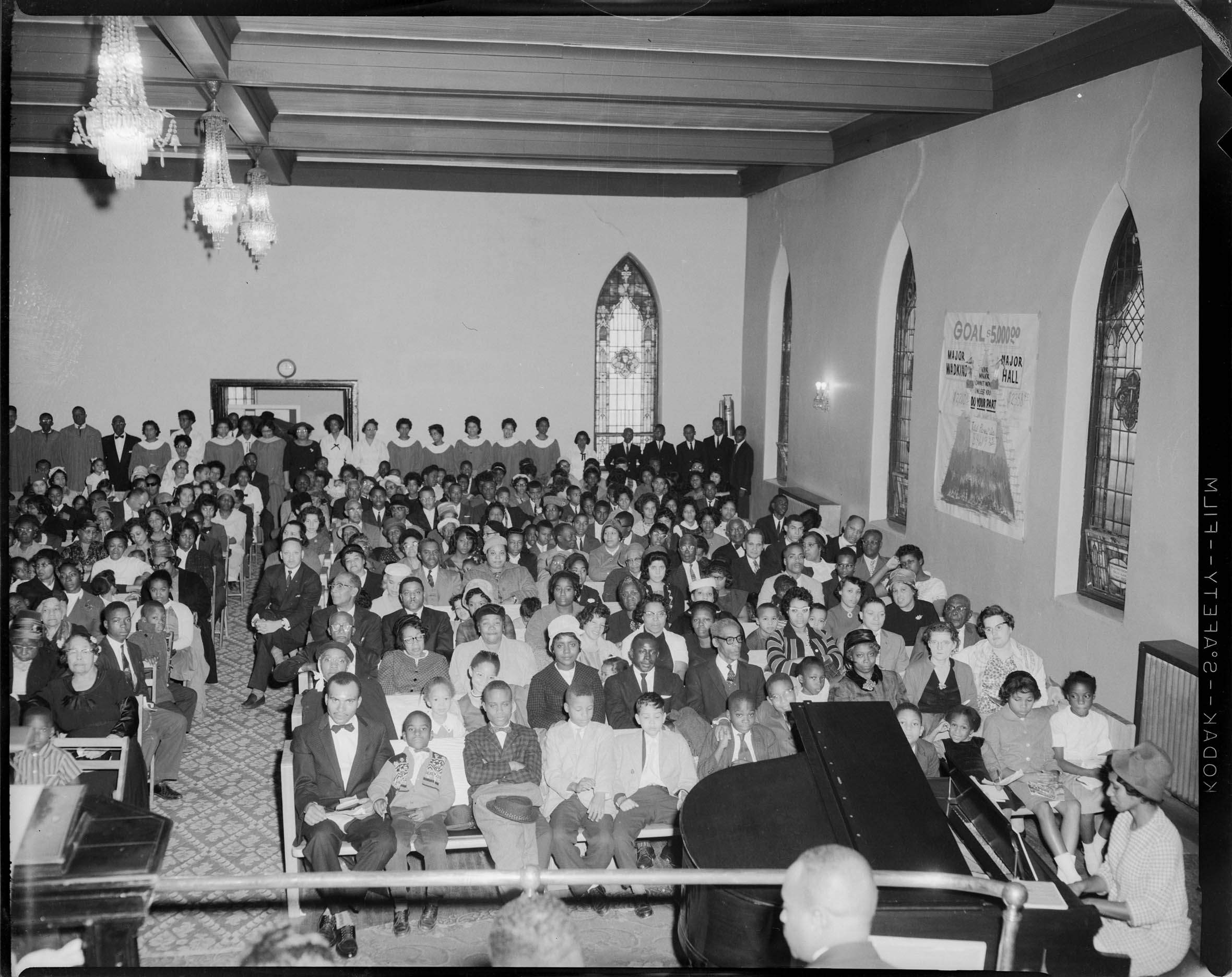 Congregation of women and men gathered in small church with choir standing in background Congregation of women and men gathered in small church with choir standing in background