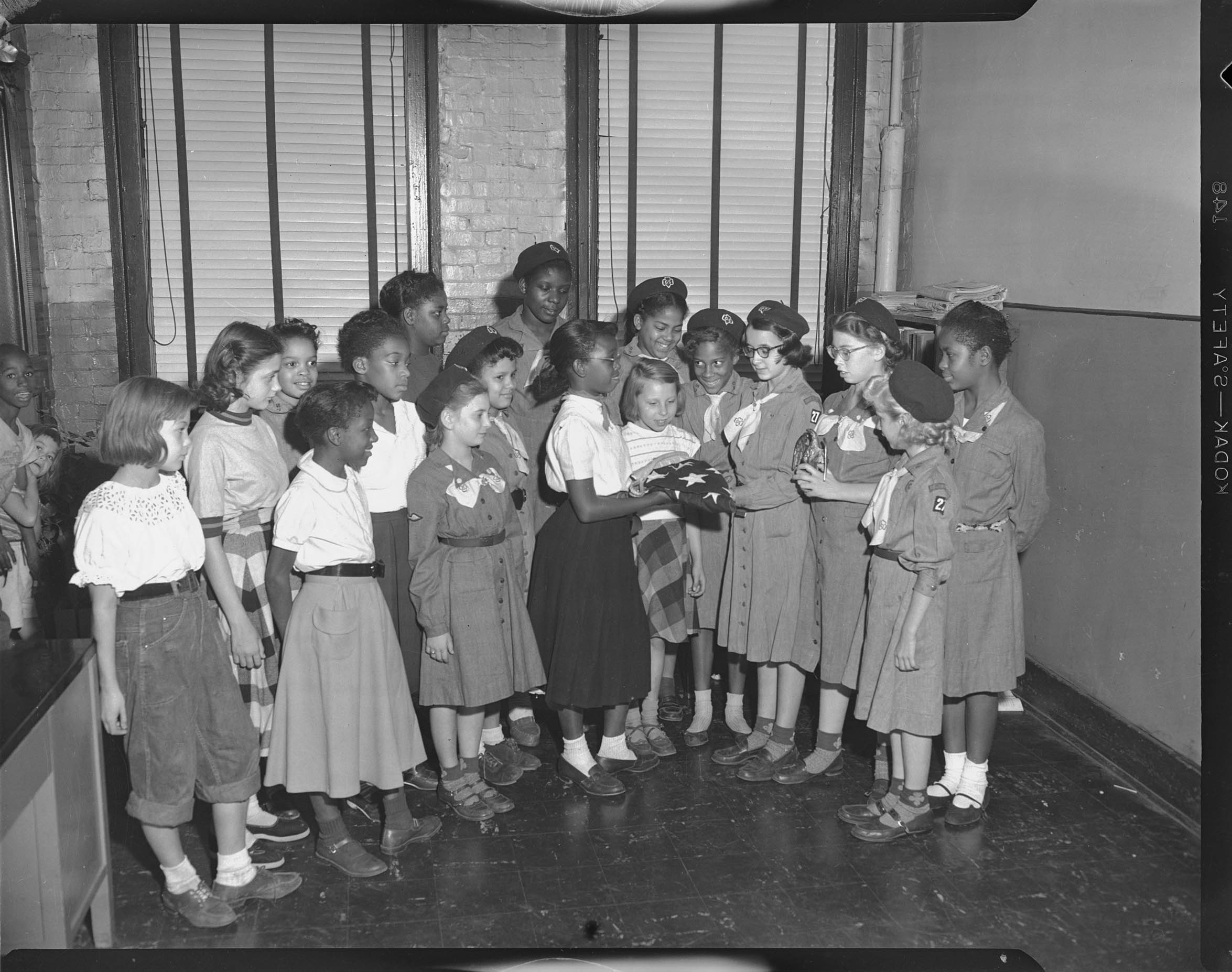 Group Portrait Of Seventeen Girls Standing Including One Center Right Wearing Girl Scout Uniform Hat Eyeglasses Light Colored Neck Scarf And Dress With Troop 27 Patch On Left Sleeve Holding Folded American