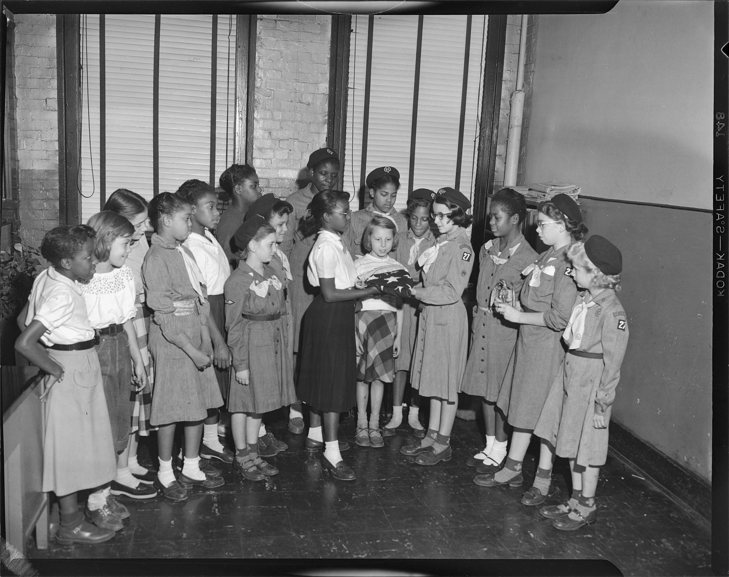 Group Portrait Of Seventeen Girls Standing Including One Center Right Wearing Girl Scout Uniform Hat Eyeglasses Light Colored Neck Scarf And Dress With Troop 27 Patch On Left Sleeve Holding Folded American