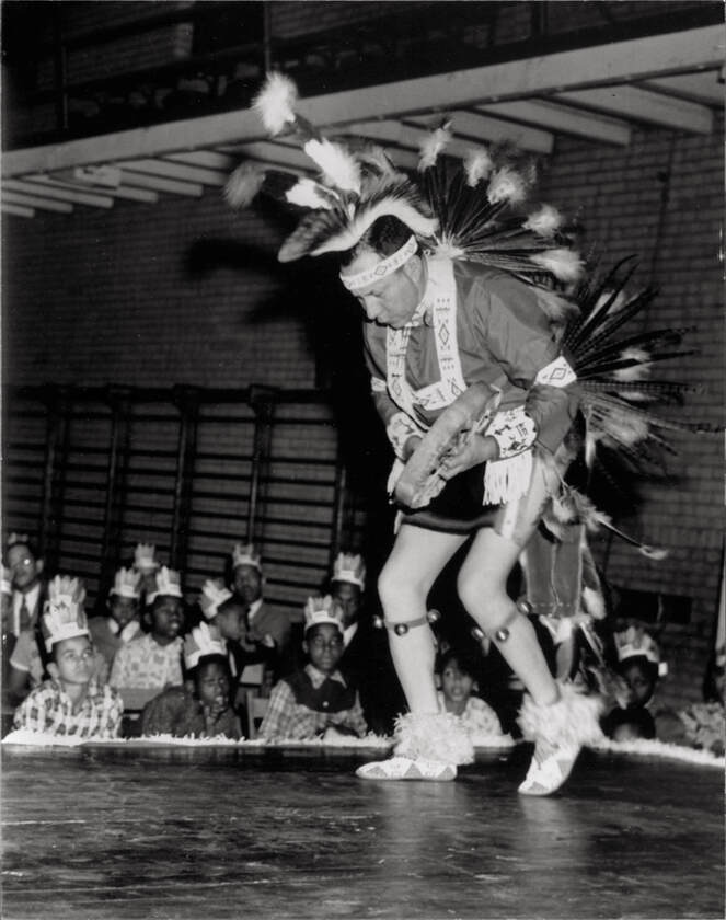Man wearing Native American dress performing dance to audience of